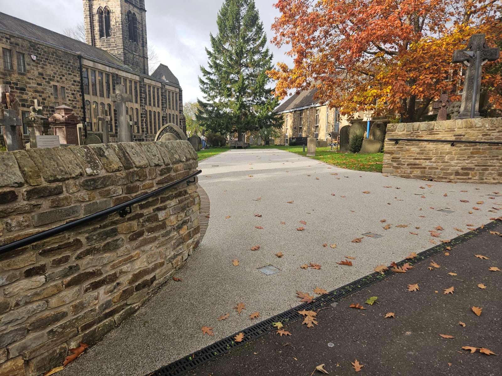 Completed light-coloured resin-bound access route through a churchyard, bordered by stone walls, headstones and mature trees.
