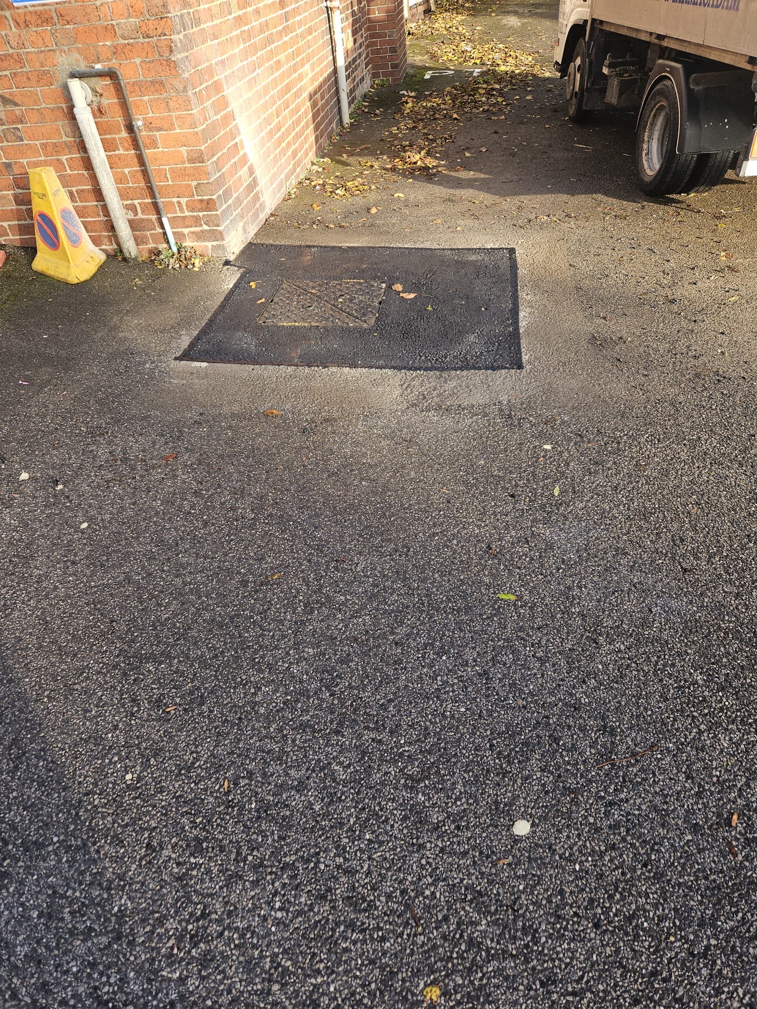 Tarmac repair around a manhole cover, reinstated flush with the surrounding asphalt surface beside a brick building.