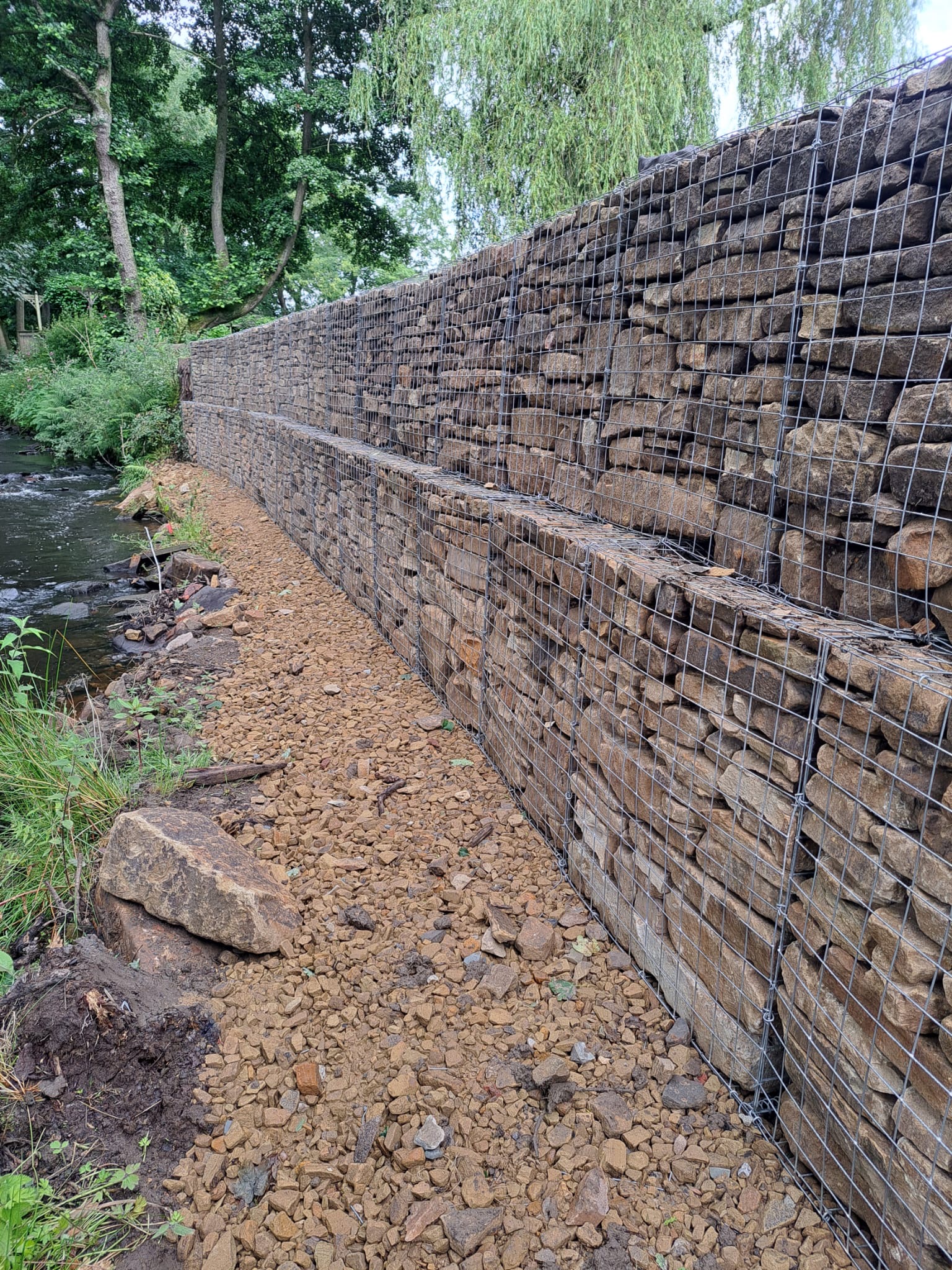 Stone-filled gabion retaining wall installed along a riverbank, providing erosion control and structural support in a natural setting.